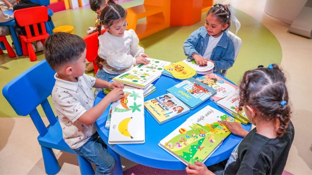 Four young children sitting around a blue table reading colourful picture books in a brightly coloured reading room