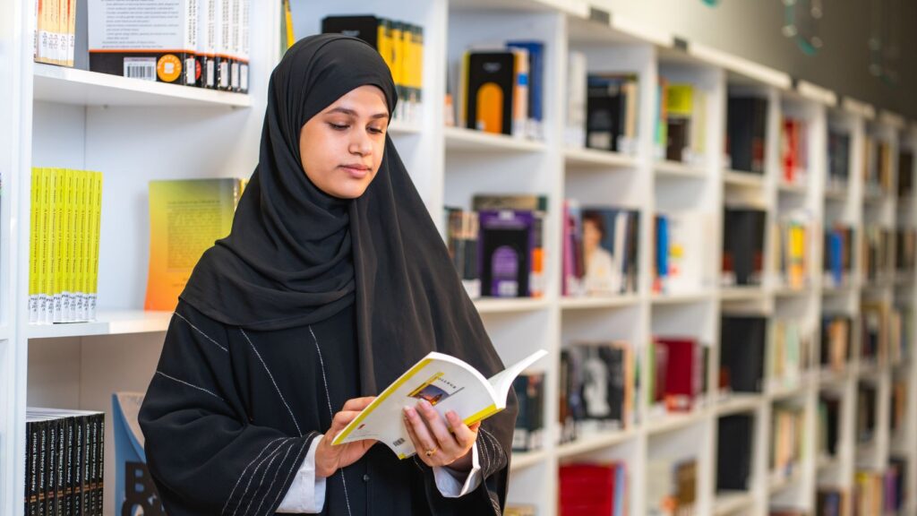 A young woman wearing a hijab reading a book in a college library surrounded by bookshelves